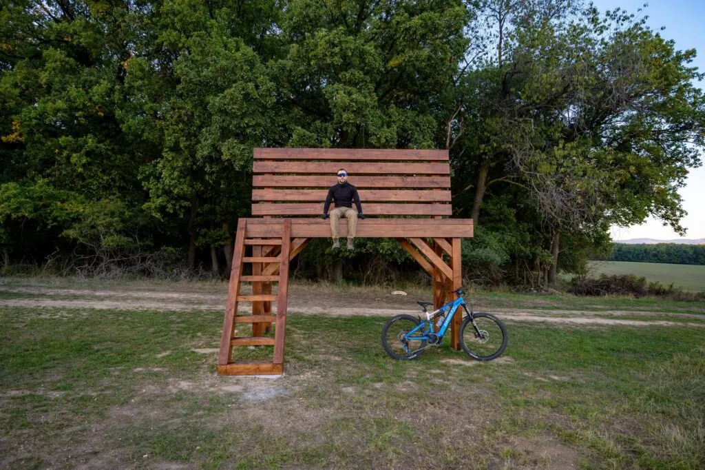 Veľká drevená lavička pri Brezolupoch s návštevníkom a bicyklom pred okrajom lesa.