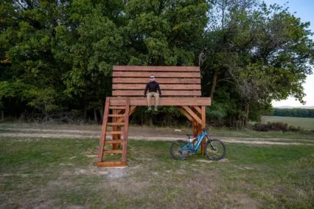 Veľká drevená lavička pri Brezolupoch s návštevníkom a bicyklom pred okrajom lesa.
