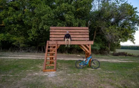 Veľká drevená lavička pri Brezolupoch s návštevníkom a bicyklom pred okrajom lesa.